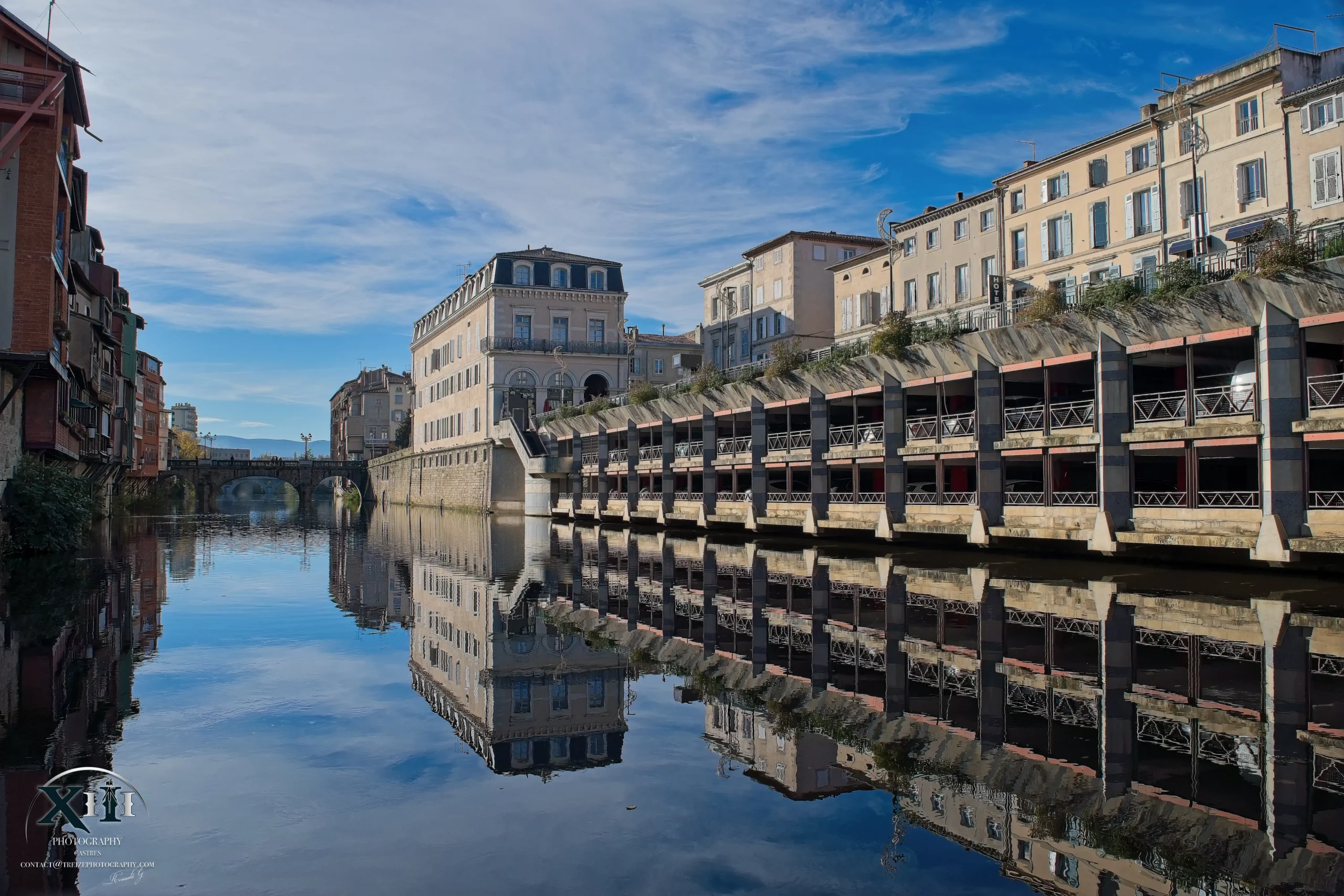 Rivière-Agout-Arcades-Castres-Tarn.webp Vue pittoresque de la rivière Agout de la ville de Castres, Tarn, avec les arcades emblématiques et les bâtiments historiques reflétés dans l’eau sous un ciel bleu.