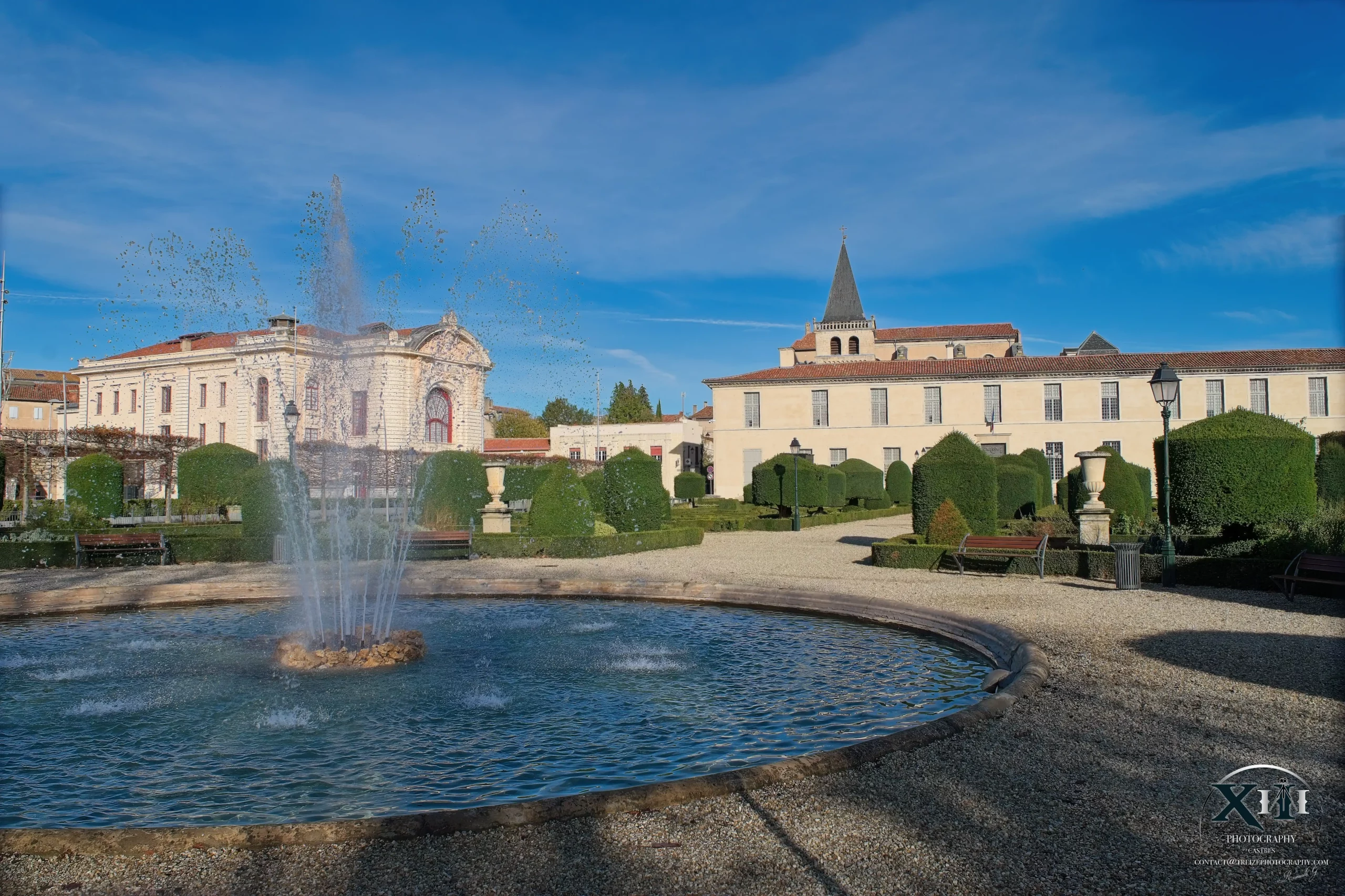 jardin-eveche-fontaine-theatre-musee-goya-castres.webp Fontaine du jardin de l’Évêché devant le théâtre municipal et le musée Goya de la ville de Castres, sous un ciel bleu.